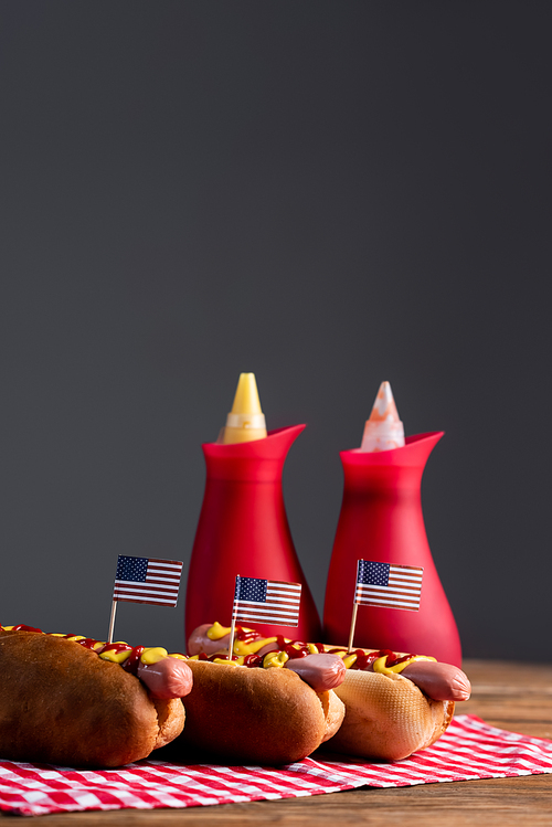 blurred sauce bottles and hot dogs with small american flags on plaid table napkin isolated on grey