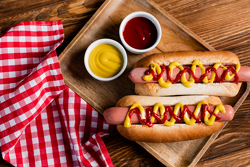 top view of hot dogs, ketchup and mustard near plaid napkin on wooden table