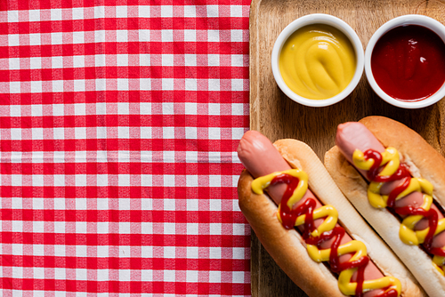 top view of wooden tray with hot dogs and bowls with mustard and ketchup on plaid tablecloth