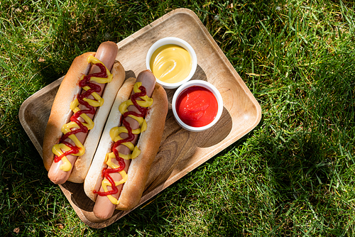 top view of wooden tray with hot dogs, mustard and ketchup on green grass