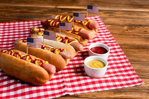 tasty hot dogs with small american flags near sauces and checkered napkin on wooden table