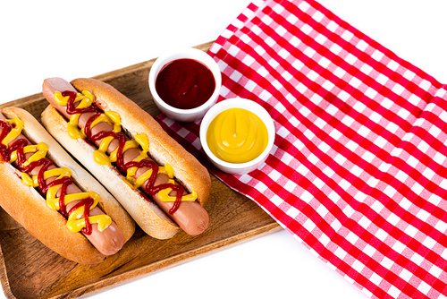 wooden tray with hot dogs, mustard and ketchup near plaid table napkin isolated on white