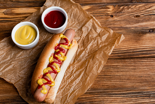 delicious hot dog near bowls with sauces and crumpled kraft paper on wooden table