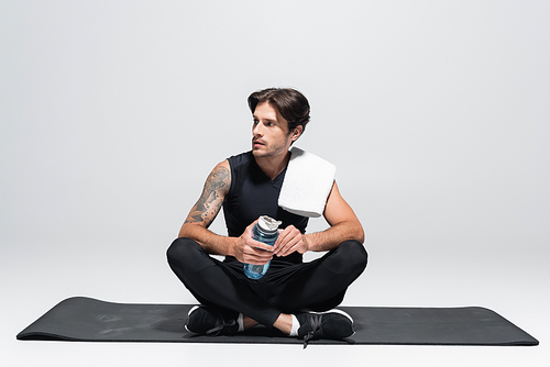 Brunette sportsman with towel and sports bottle sitting on fitness mat on grey background