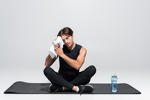 Sportsman wiping face with towel near sports bottle on fitness mat on grey background