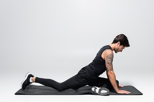 Side view of sportsman stretching leg on fitness mat on grey background