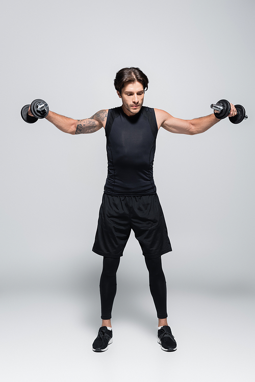 Full length of tattooed sportsman training with dumbbells on grey background