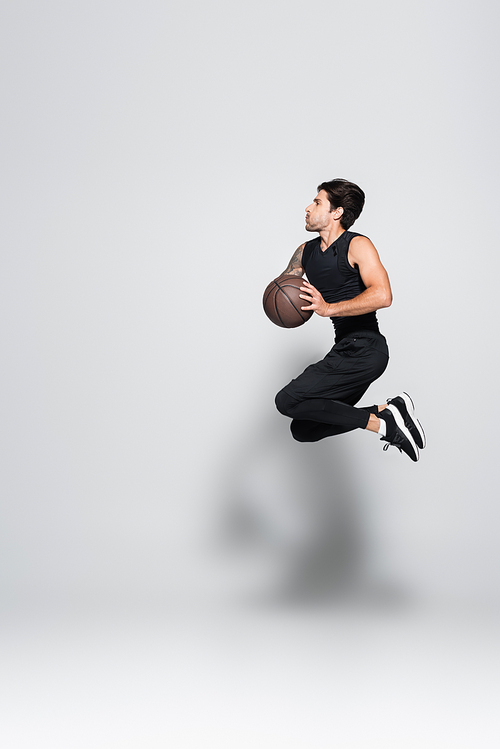Side view of tattooed sportsman with basketball ball jumping on grey background