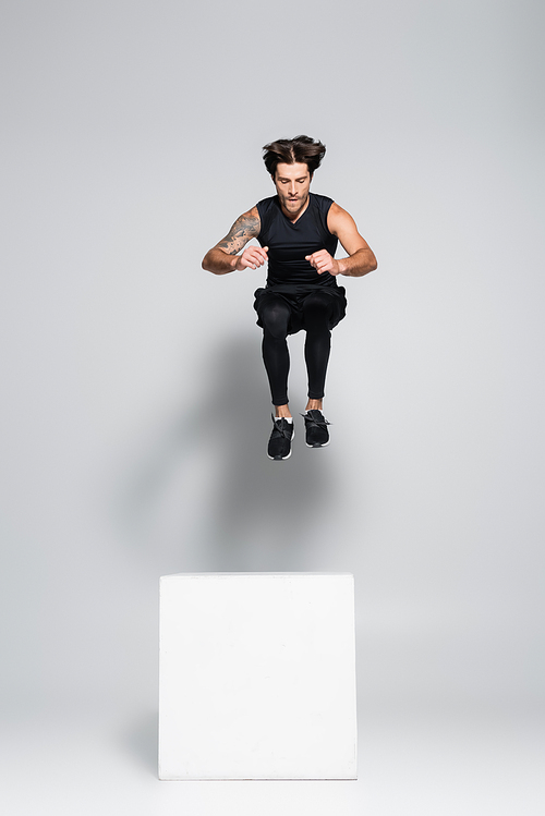 Athletic sportsman jumping above cube on grey background