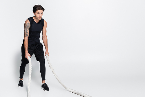 Tattooed sportsman training with battle ropes on grey background with copy space