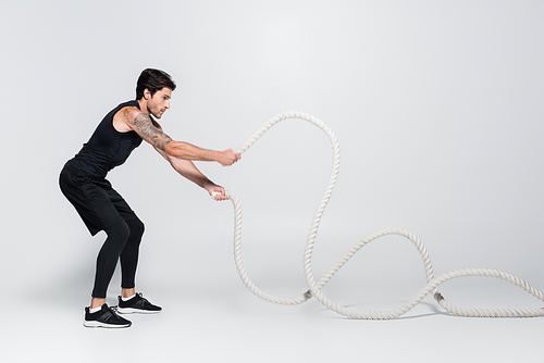 Side view of sportsman working out with battle ropes on grey background