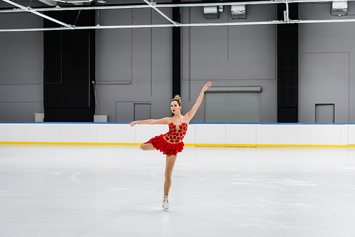 full length of figure skater in red dress performing camel spin in ice arena