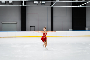 full length of young woman in dress figure skating with outstretched hands in professional ice arena
