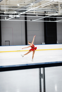 full length of happy woman in red dress figure skating with outstretched hands in professional ice arena