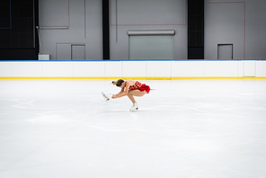 full length of young woman in dress performing sit spin in professional ice arena