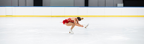 full length of young woman in dress performing sit spin in professional ice arena, banner