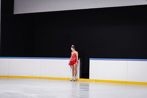 full length of young worried figure skater in red dress standing on ice rink