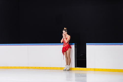 full length of worried figure skater in red dress standing on ice rink