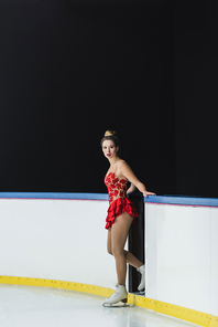 full length of young worried figure skater in red dress standing near ice arena