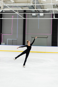 full length of young woman in black bodysuit gesturing while skating in professional ice arena