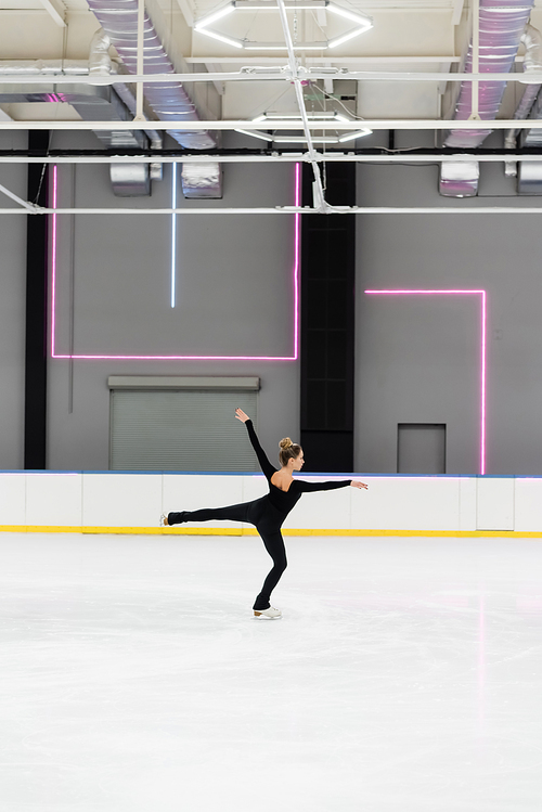 full length of young woman in black bodysuit with outstretched hands figure skating in professional ice arena