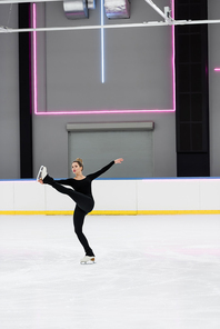 full length of figure skater in black bodysuit skating with outstretched hand in professional ice arena