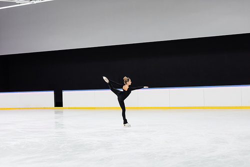 side view of figure skater in black bodysuit skating with outstretched hand in professional ice arena