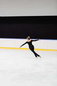 full length of professional figure skater in black bodysuit skating with outstretched hands in ice arena
