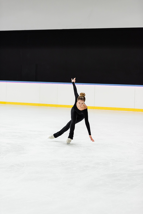 full length of young professional figure skater in black bodysuit skating with outstretched hand in ice arena