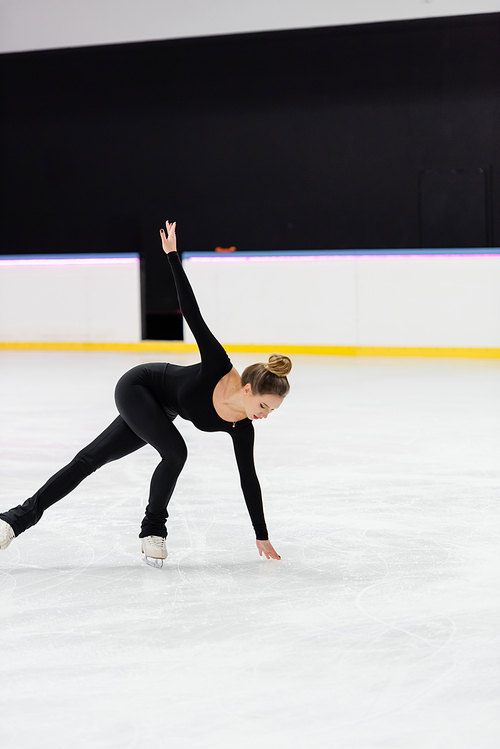 full length of professional figure skater in black bodysuit skating with outstretched hand and touching frozen ice in arena