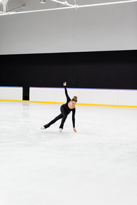 full length of young figure skater in black bodysuit skating with outstretched hand and touching frozen ice in arena