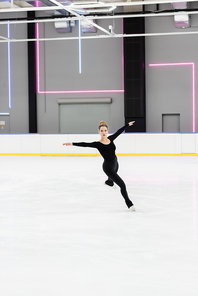 full length of professional figure skater in black bodysuit skating with outstretched hands in ice arena with neon lights