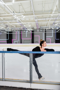 young woman in white figure skates stretching near ice rink