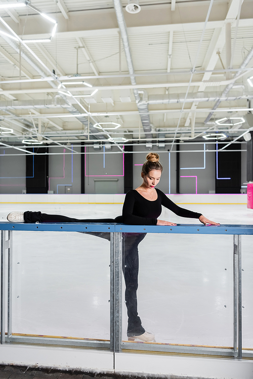young woman in white figure skates stretching near ice arena