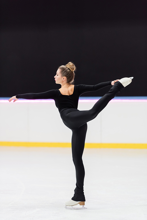 side view of professional figure skater in black bodysuit stretching with outstretched hand in ice arena