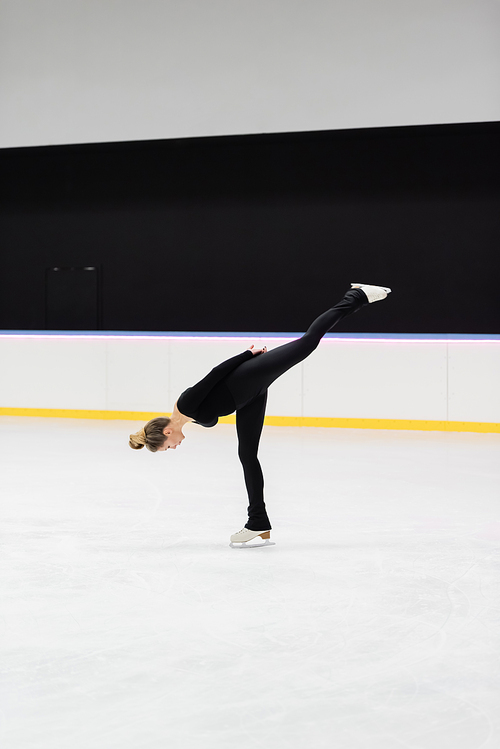 full length of professional figure skater in black bodysuit skating while bending in ice arena