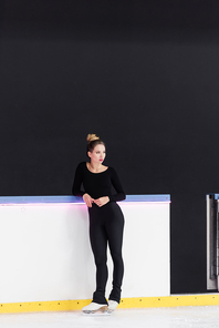 full length of young figure skater in black bodysuit standing on ice skates near frozen ice arena