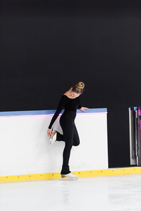 full length of young figure skater in black bodysuit checking blade on ice skates near frozen ice arena