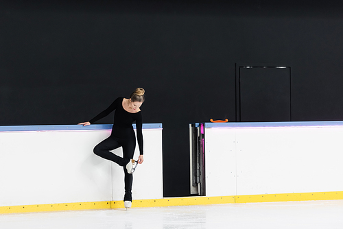 full length of figure skater in black bodysuit checking blade on ice skates near frozen ice arena
