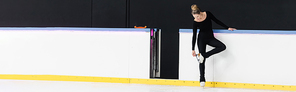full length of figure skater in black bodysuit checking blade on ice skates near frozen ice arena, banner