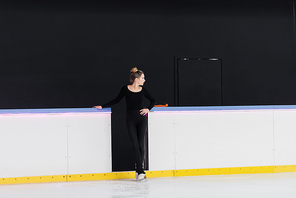full length of young figure skater in black bodysuit standing with hand on hip near frozen ice arena