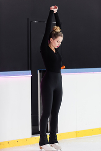 full length of young figure skater in black bodysuit and ice skates stretching near frozen ice arena