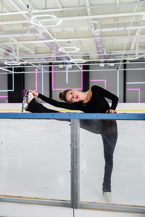full length of professional figure skater in black bodysuit and ice skates stretching near frozen ice arena