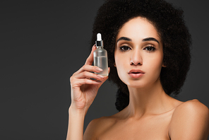 pretty african american woman with bare shoulders holding bottle of cosmetic serum isolated on grey