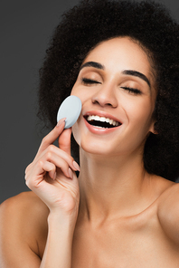 happy african american woman with closed eyes holding cosmetic sponge isolated on grey