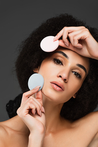 young african american woman with perfect skin and natural makeup posing with cosmetic sponges isolated on grey