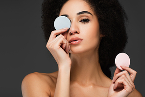 african american woman with naked shoulders obscure eye with cosmetic sponge isolated on grey