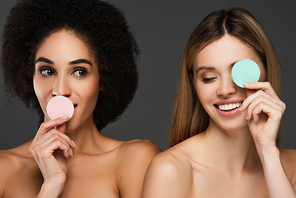 cheerful multiethnic women with bare shoulders holding cosmetic sponges near faces isolated on grey
