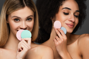 pretty interracial women with natural makeup posing with round cosmetic sponges isolated on grey