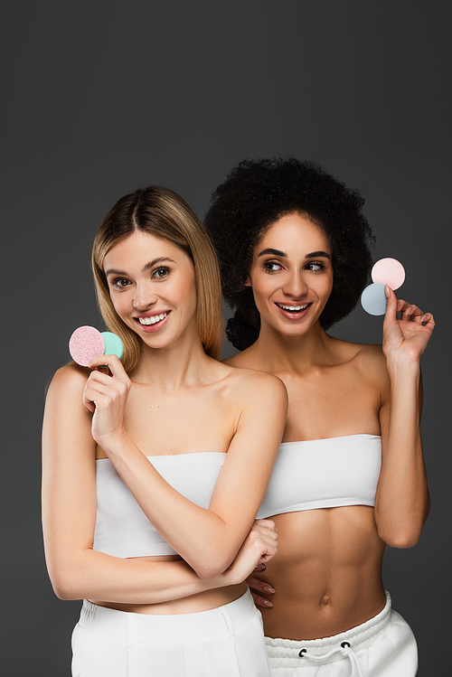 young and slim multiethnic women posing with cosmetic sponges isolated on grey